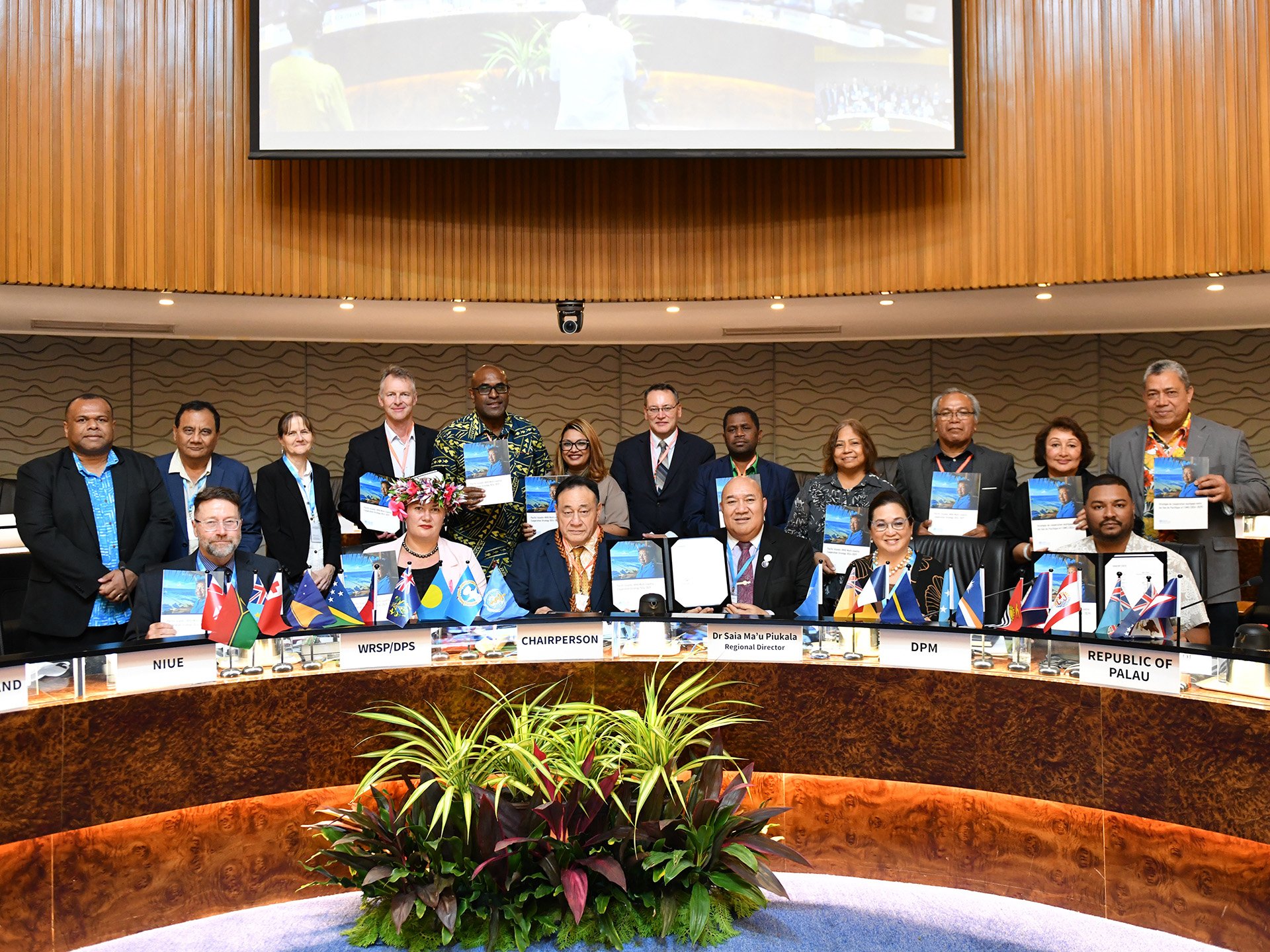People stand behind a conference table, holding documents at an international event. The table has nameplates for different countries and designations with small flags and a large screen background projection.